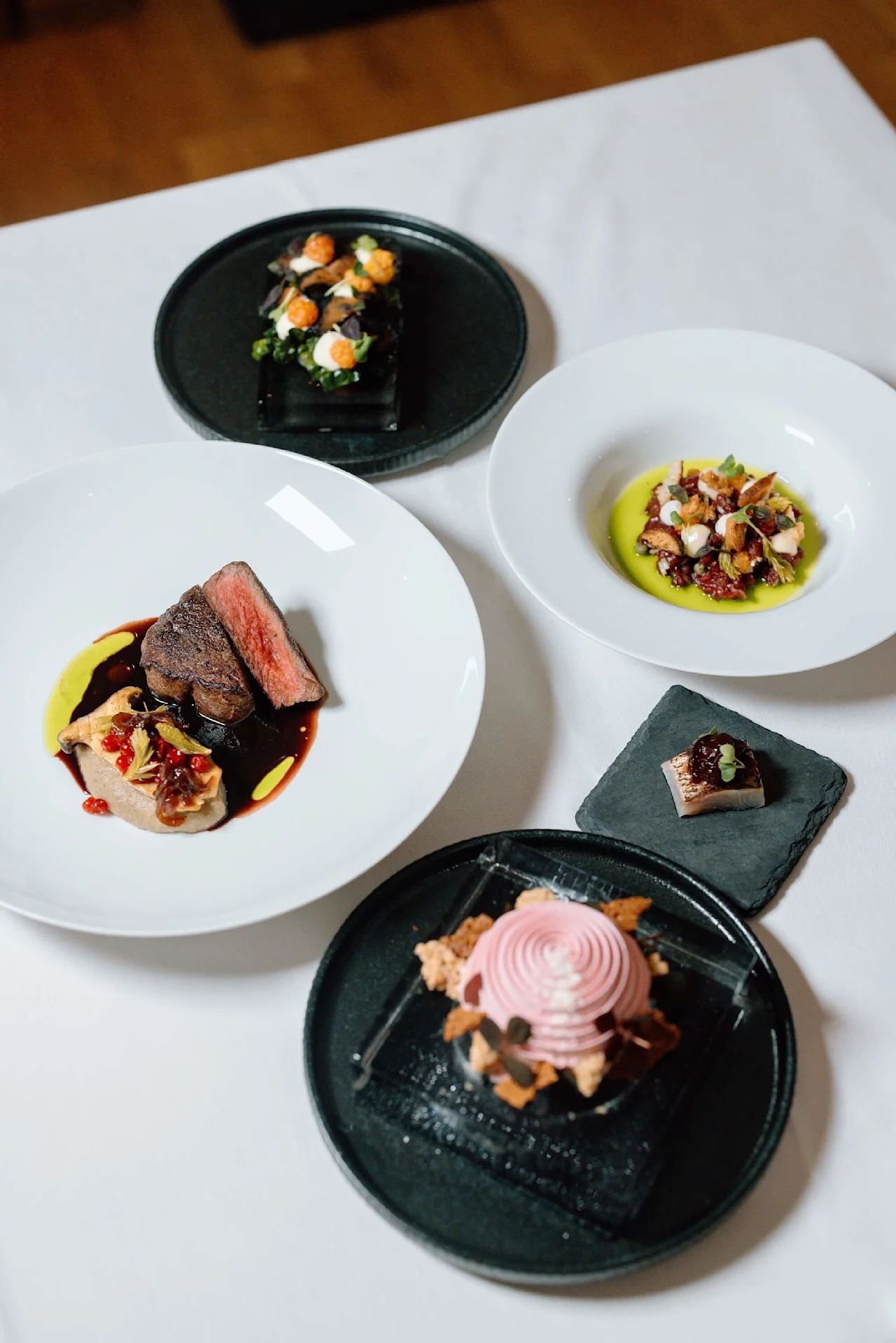 Et udvalg af gourmet retter præsenteret på elegante tallerkener, med kød, grøntsager og en lyserød dessert, arrangeret på en hvid dug.