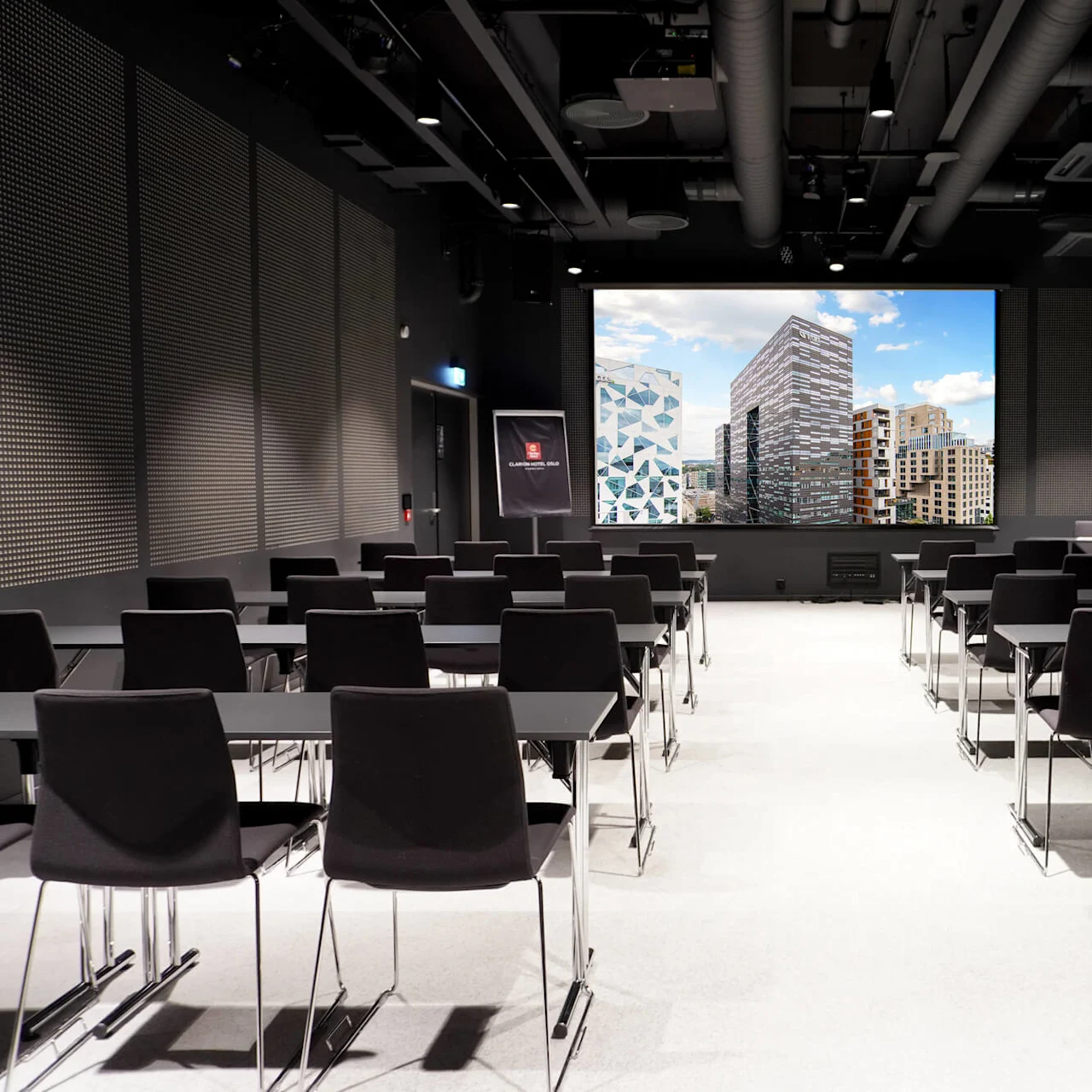 A conference room with rows of black chairs facing a large screen showing modern buildings. A black sign reads "CLARION SIGN." The room has dim lighting and minimalist design.