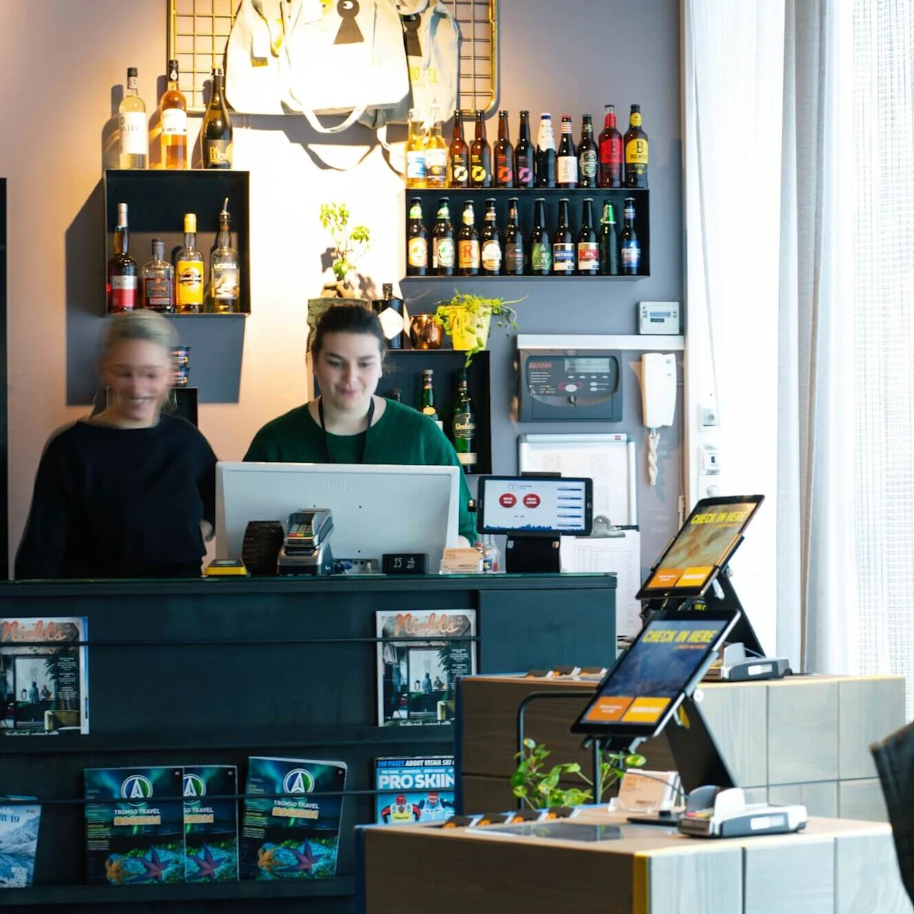 Two people stand behind a counter, interacting with a computer. Shelves of bottles and a plant decorate the background in a cozy, well-lit room with curtains.