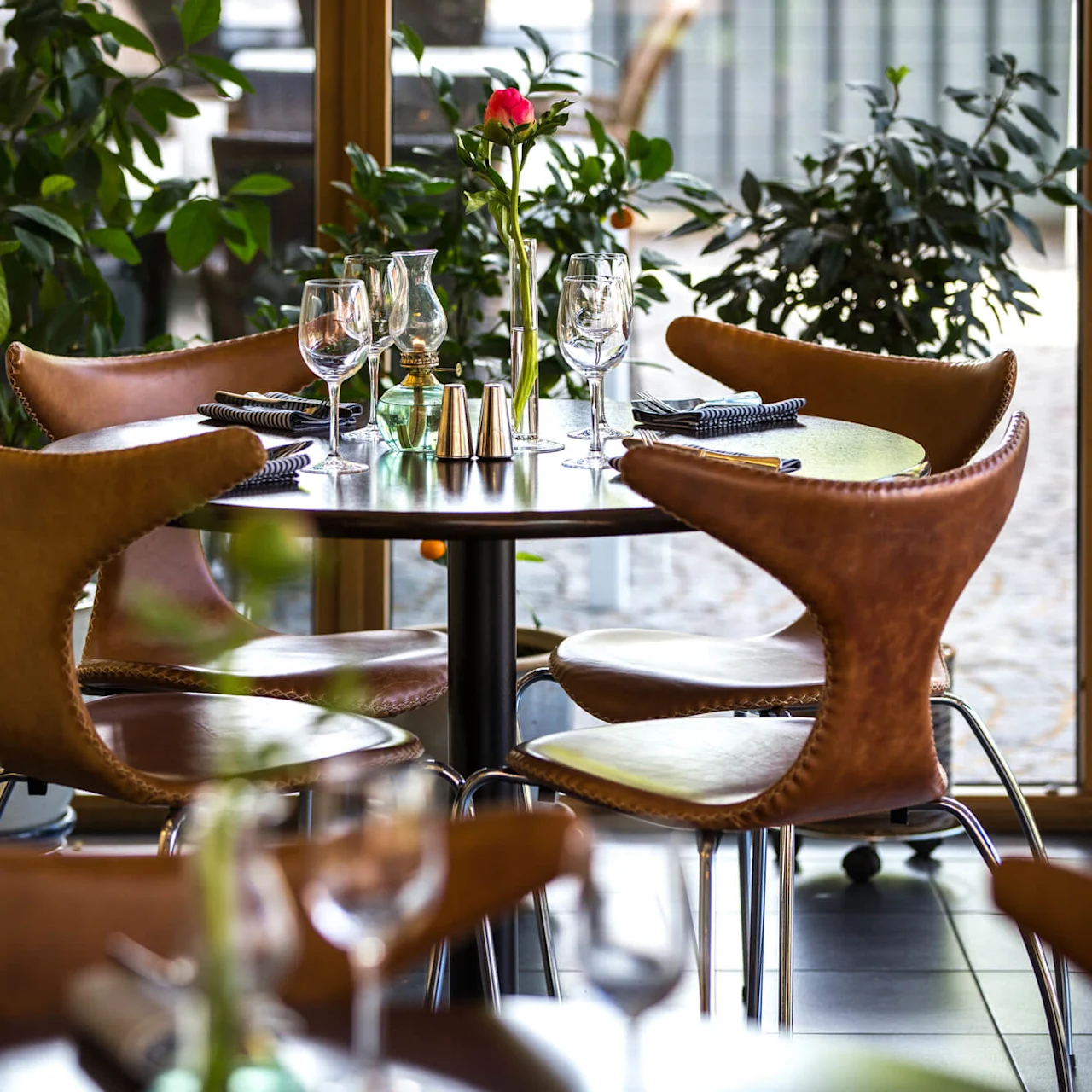 Elegant dining setup with empty wine glasses on a round table, surrounded by plush chairs, near a window with indoor plants.