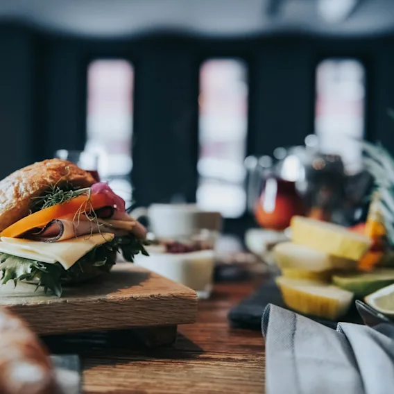 En smörgås med ost, sallad och skivade grönsaker ligger på en träplatta. Runt omkring finns tropiska frukter, en croissant och porslin i en svagt upplyst matsal.