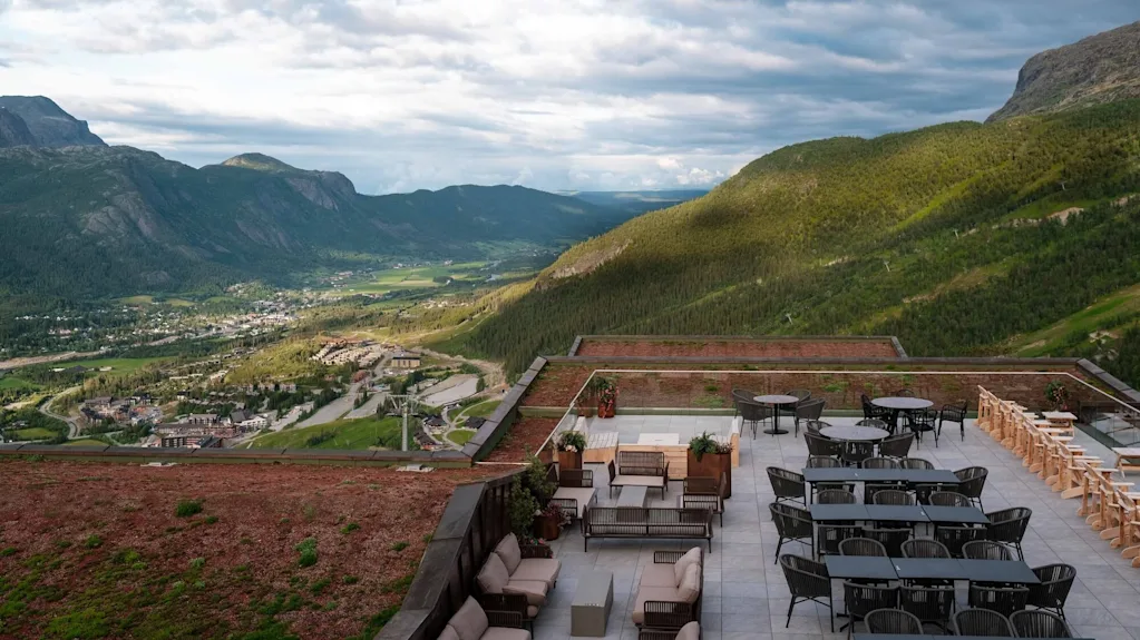 Tagterrasse med moderne udendørsmøbler, står stille, med udsigt over en grøn dal med fjerne bjerge og en lille by nedenfor.
