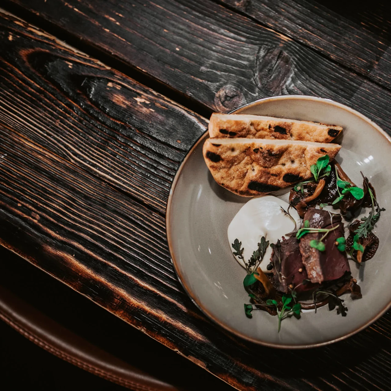 A ceramic plate containing sliced meat, greens, and flatbread is placed on a rustic wooden table. There is also a dollop of white sauce on the plate.