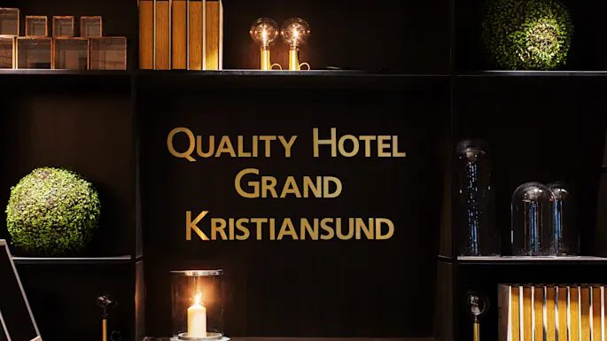 Quality Hotel Grand Kristiansund, text in gold letters, decorates a dark shelf. Books, round green plants, two glass-covered bulbs, and a candle are arranged on surrounding shelves.