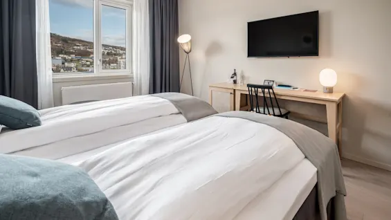 To enkeltsenge med hvidt sengetøj i et pænt hotelværelse, med skrivebord, TV og stort vindue med udsigt.