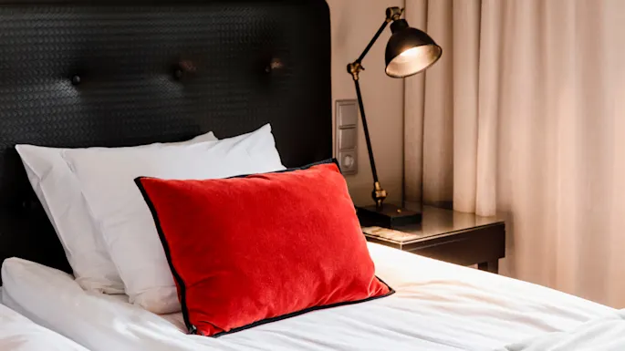 Seng med en rød pute hviler på hvite laken, ledsaget av en svart lampe på et sidebord, omgitt av beige gardiner.