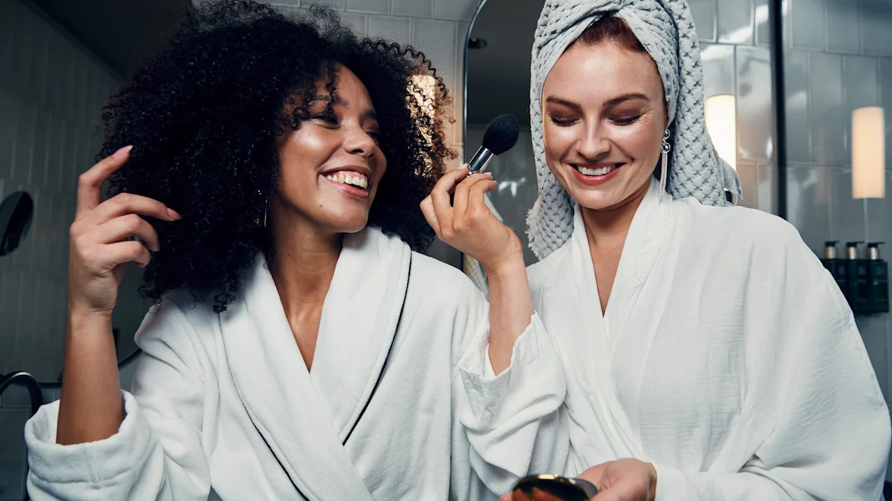 To venner i badekåper deler et gledelig øyeblikk mens den ene sminker den andre på et godt opplyst bad.
