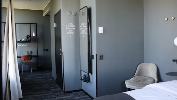 A modern hotel room with a bed, a desk area, and a mirror. Text on the wall reads IF YOU CANT FIND THE BUTLER DO IT YOURSELF.