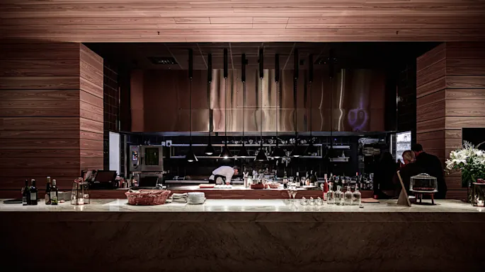 A chef works in a modern, open kitchen with wooden accents and sleek equipment. People dine nearby, while bottles and tableware sit on the counter, creating a cozy atmosphere.