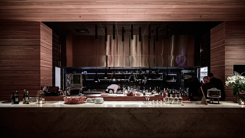 A chef works in a modern, open kitchen with wooden accents and sleek equipment. People dine nearby, while bottles and tableware sit on the counter, creating a cozy atmosphere.