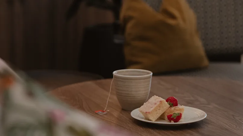 A cup of tea sits on a wooden table beside a plate with cake and strawberries, in a cozy room with cushions.