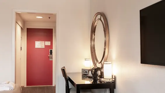 Viihtyisä hotellihuone, jossa on työpöytä, peili, lamput ja vedenkeitin, valmiina vieraalle rentoutumaan tai työskentelemään. Punainen ovi johtaa tilaan.