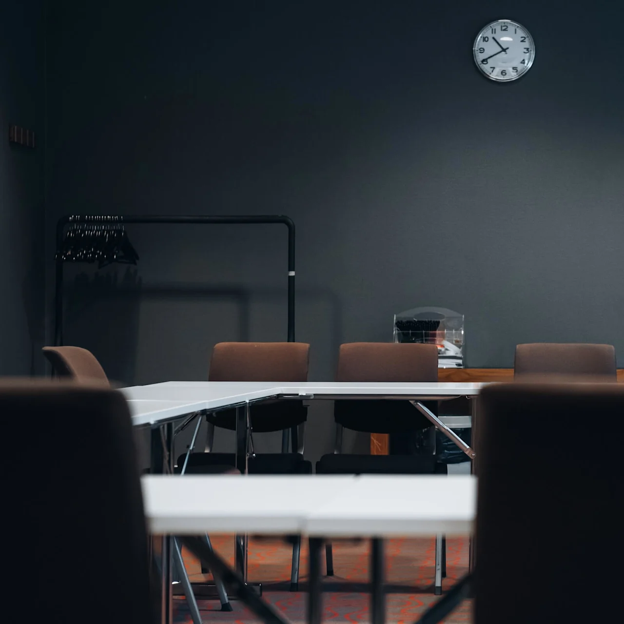 Meeting room with white tables and brown chairs arranged in a U-shape. A wall clock shows 12:15. Coatrack on the left, framed pictures on the right wall.