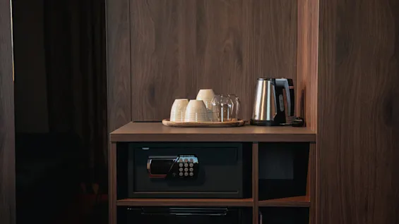 Et koselig hotellromoppsett med en vannkoker, kopper og glass på et trebrett, sammen med et sikkert safe for personlige eiendeler.