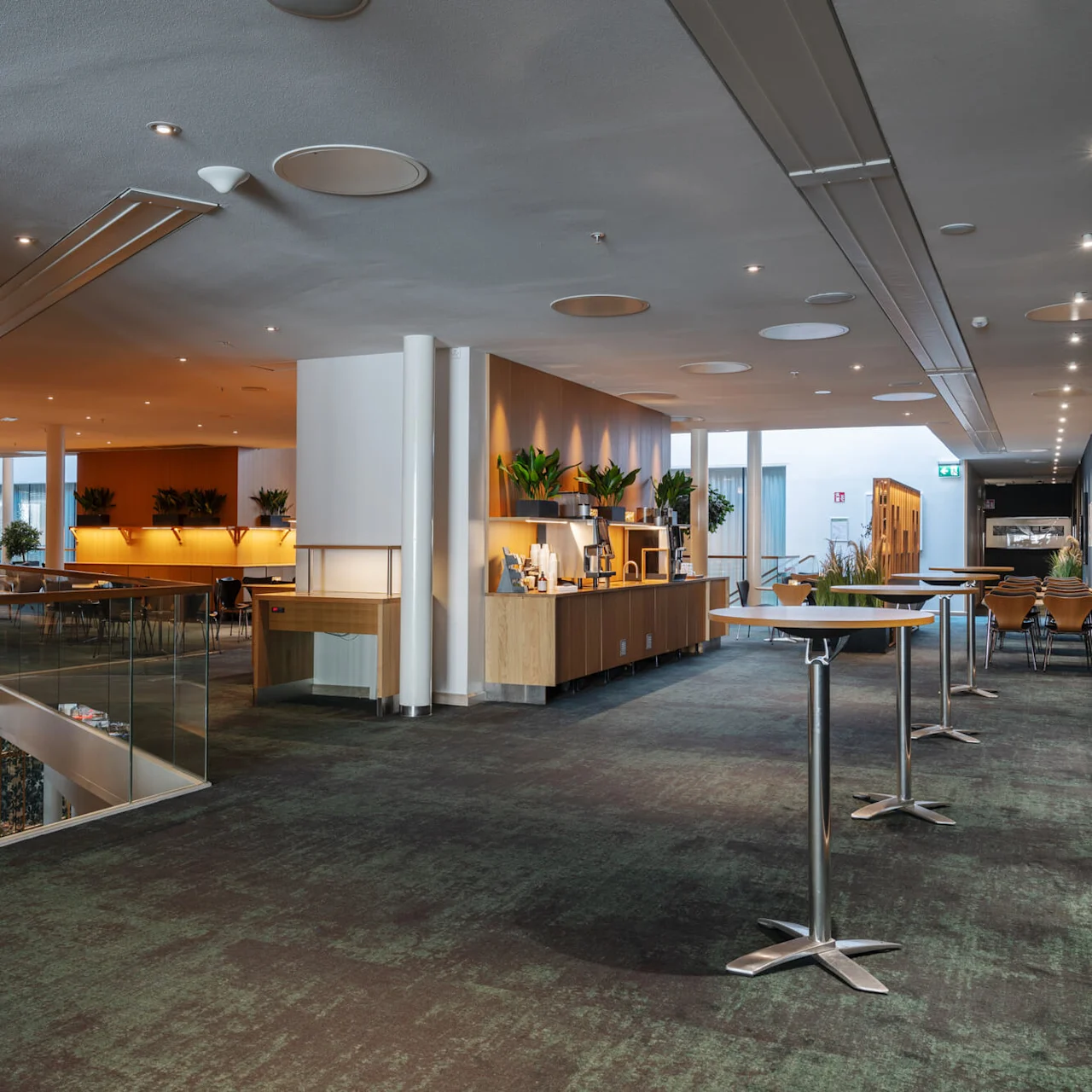 Modern loungeområde med bord och stolar arrangerade i en bred korridor, omgivet av träpaneler och växter, med mjuk belysning som skapar en välkomnande atmosfär.