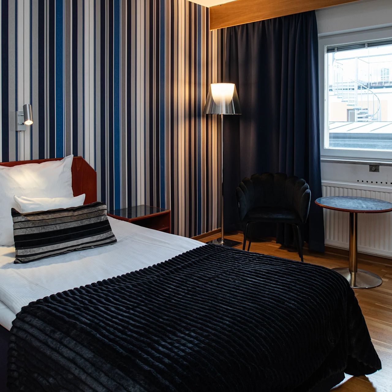 En pænt redt enkeltseng med stribet sengelinned står op ad en stribet væg. Et vindue med mørke gardiner, en lampe og et lille bord med en stol er i nærheden.