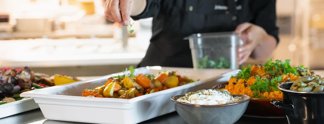 Kokki ripottelee yrttejä paahdettujen vihannesten päälle valkoiseen astiaan, ympärillä erilaisia ruokia ruostumattomasta teräksestä valmistetulla työtasolla ammattikeittiössä.