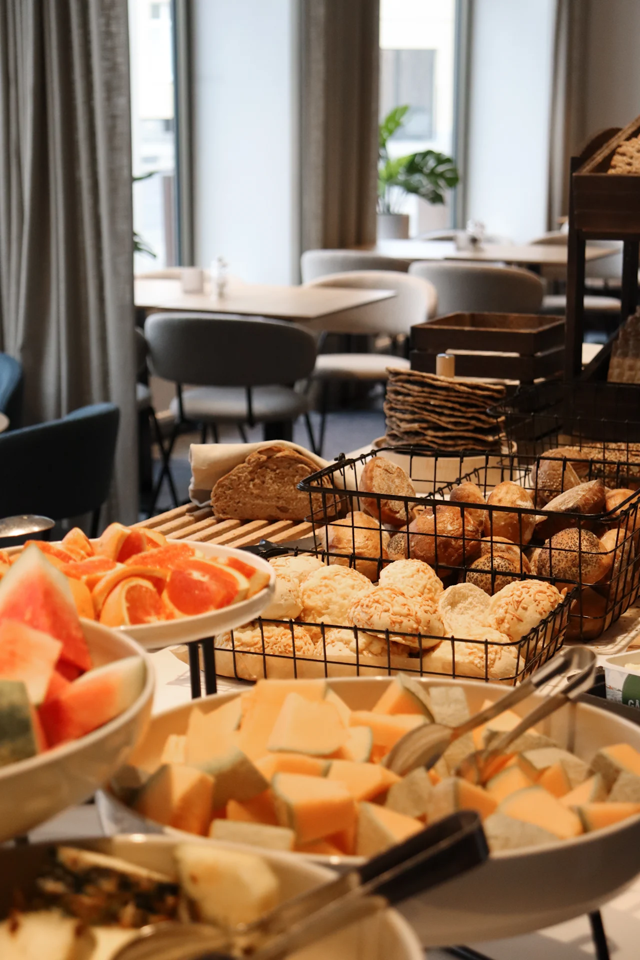 Bread and fruit are arranged on a table in a buffet setting, surrounded by dining tables and chairs in a softly lit room with large windows and green plants.