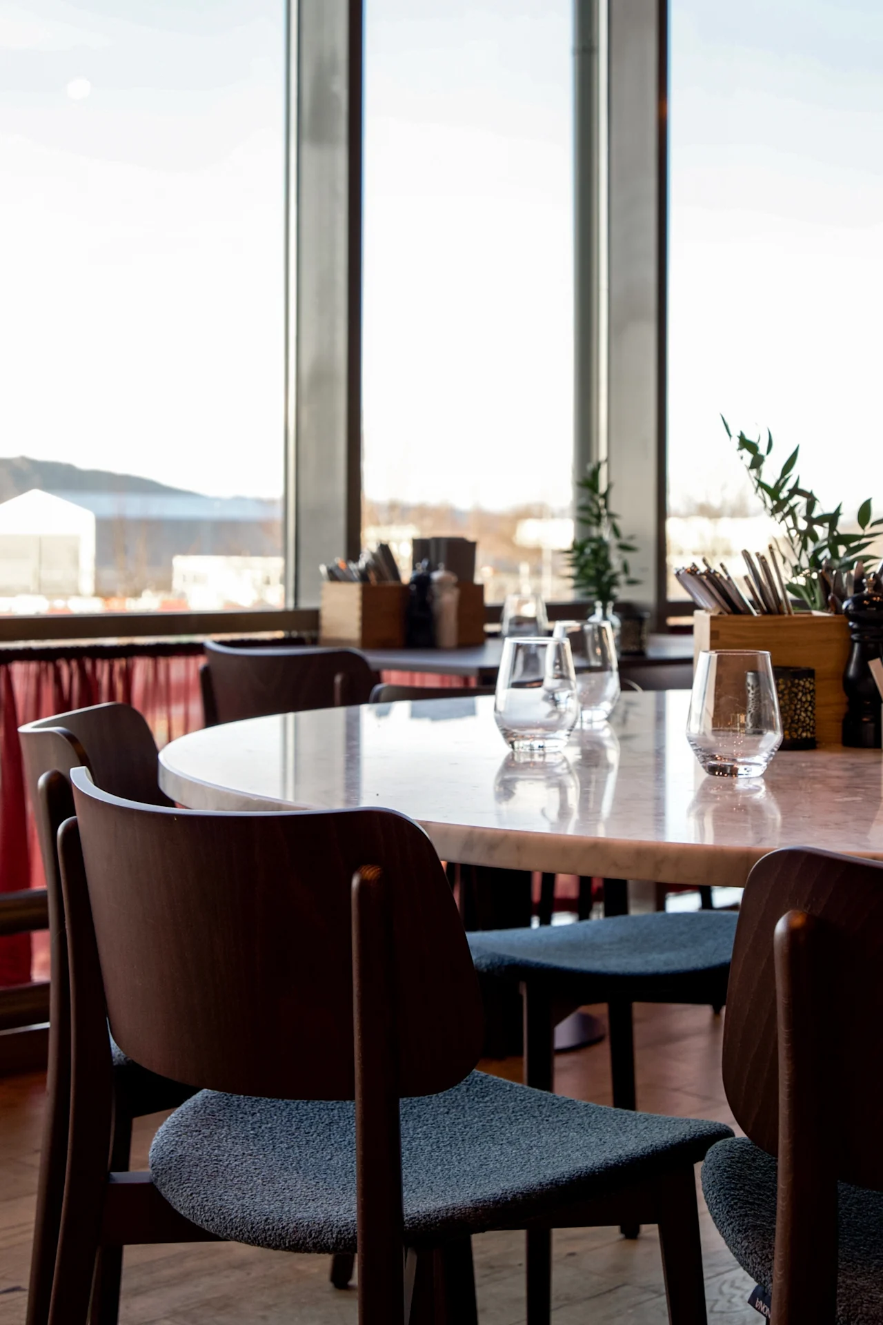 Rundt spisebord omgitt av trestoler, satt med glass. Ligger i en lys restaurant med store vinduer med utsikt over fjellene og røde gardiner under vinduene.