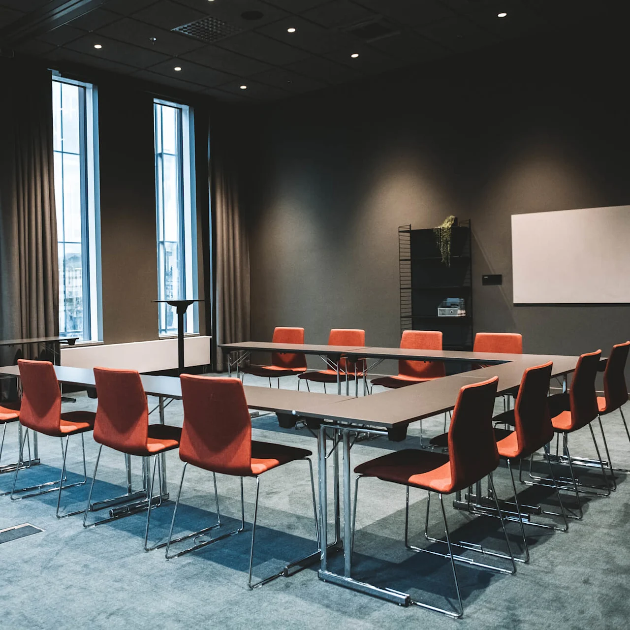 Ett U-format konferensbord med röda stolar står i ett modernt mötesrum med höga fönster, grå väggar och en blå matta. En whiteboard och hylla pryder väggen.