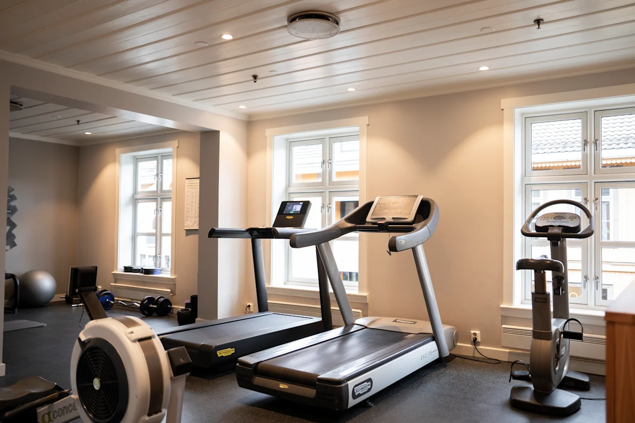 Tredemøller står ubrukte i et moderne treningsstudio, omgitt av treningsutstyr og belyst av naturlig lys gjennom store vinduer.