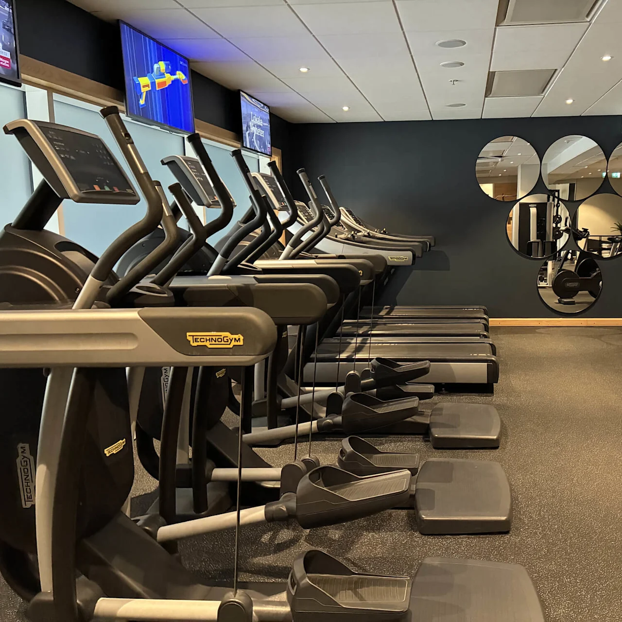 Løbebånd er opstillet på række i et moderne træningscenter, med front mod vægspejle og skærme med træningsinformation. Omgivende motionscykler bidrager til det rummelige fitnessmiljø.