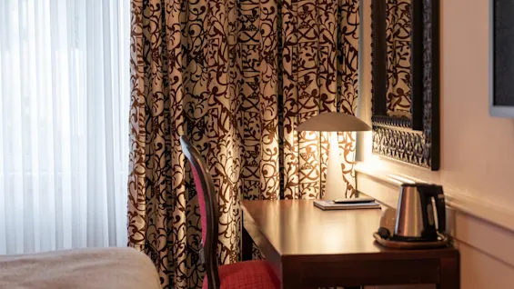 A cozy hotel room desk area with a warm lamp, a kettle, and patterned curtains at Home Hotel Grand Bodø.