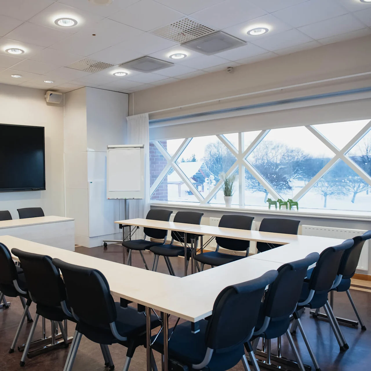 A U-shaped table arrangement with black chairs in a conference room. A large screen and flipchart stand at the front. A window reveals a snowy landscape outside.