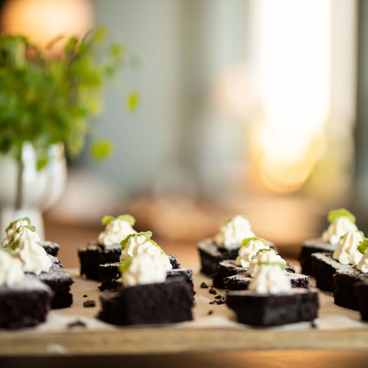 Brownie-rutor toppade med vispad grädde och myntablad vilar på bakplåtspapper. Solljus filtreras mjukt genom den suddiga bakgrunden och skapar en varm och inbjudande atmosfär.