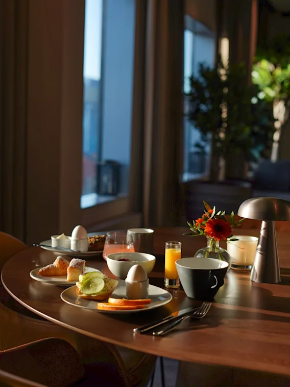 A cozy breakfast spread on a wooden table with pastries, eggs, fruit, and drinks, bathed in soft light from a large window.