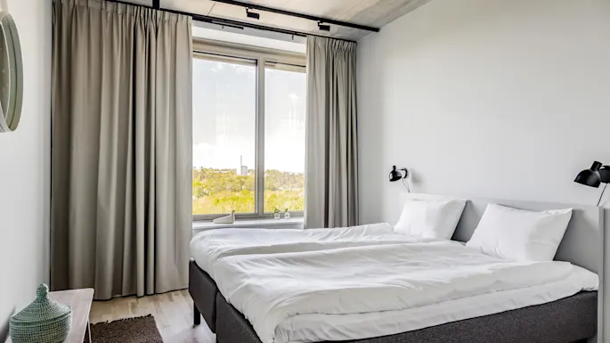 A neatly made double bed with white linens stands by a large window with light gray curtains, in a minimalistic room featuring wooden flooring and a wall-mounted lamp.