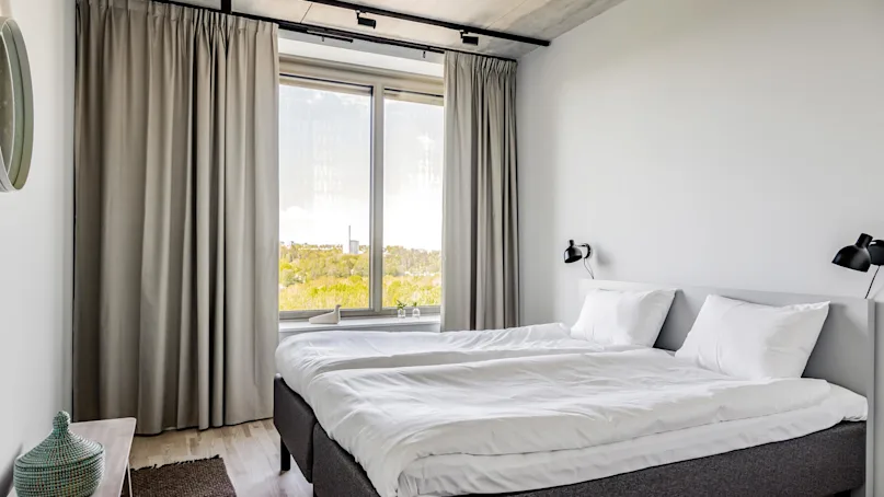 A neatly made double bed with white linens stands by a large window with light gray curtains, in a minimalistic room featuring wooden flooring and a wall-mounted lamp.