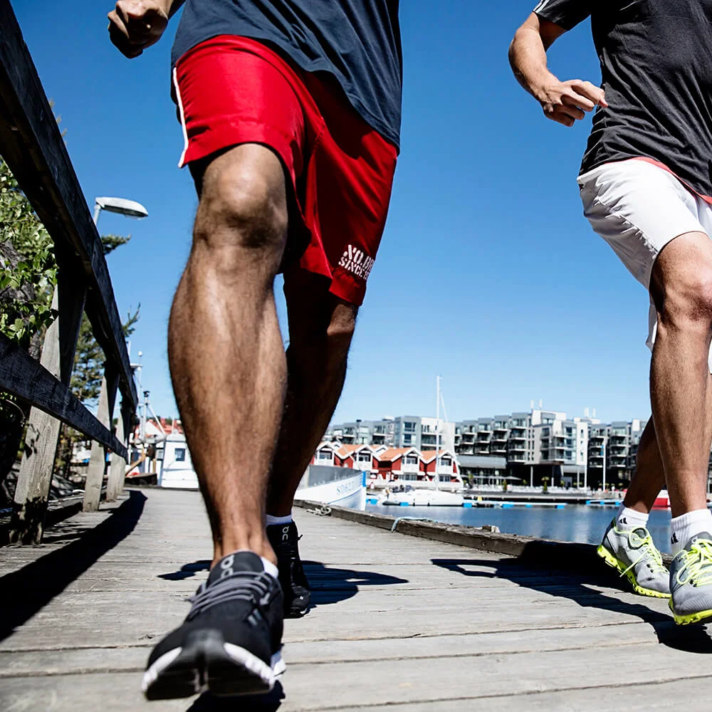 To personer jogger langs en brygge. Båter og moderne bygninger i bakgrunnen under en klar blå himmel. Tekst på shorts: "WORLD SINGLES."
