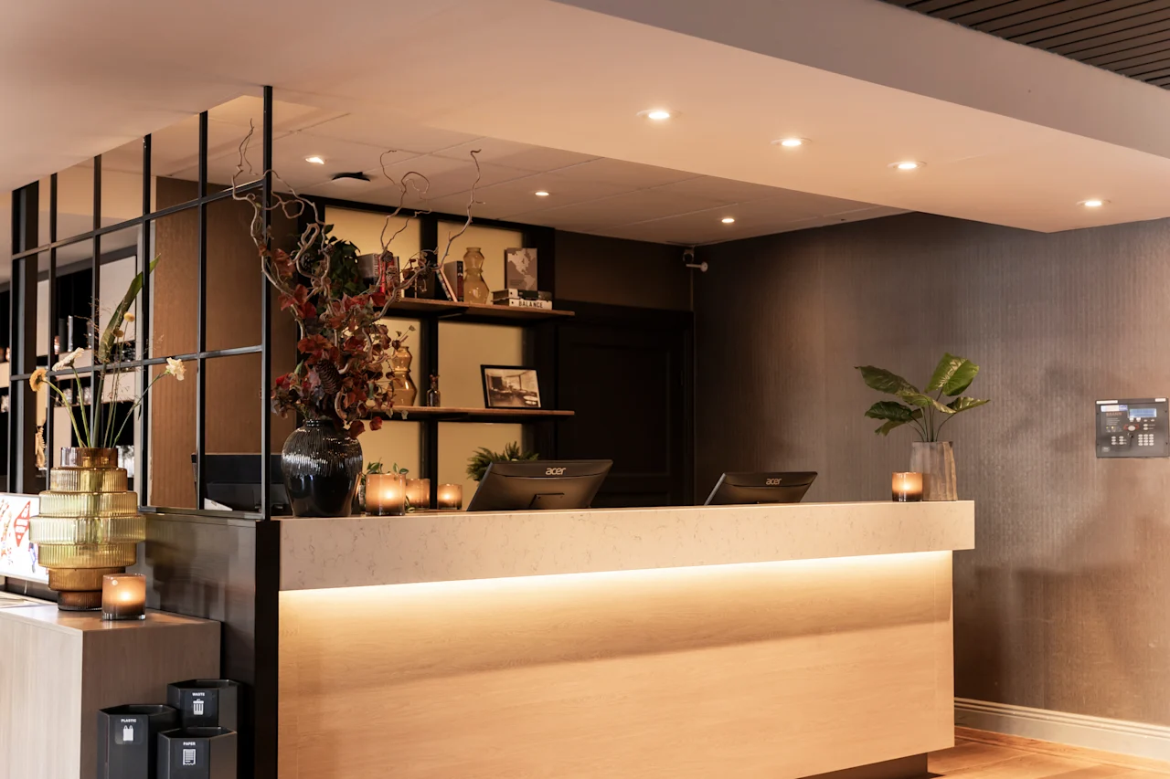 Et indbydende hotelreceptionsområde med en lyst oplyst skranke, computere og elegant indretning inklusive planter og stearinlys. Synlig etiket: PAPER.
