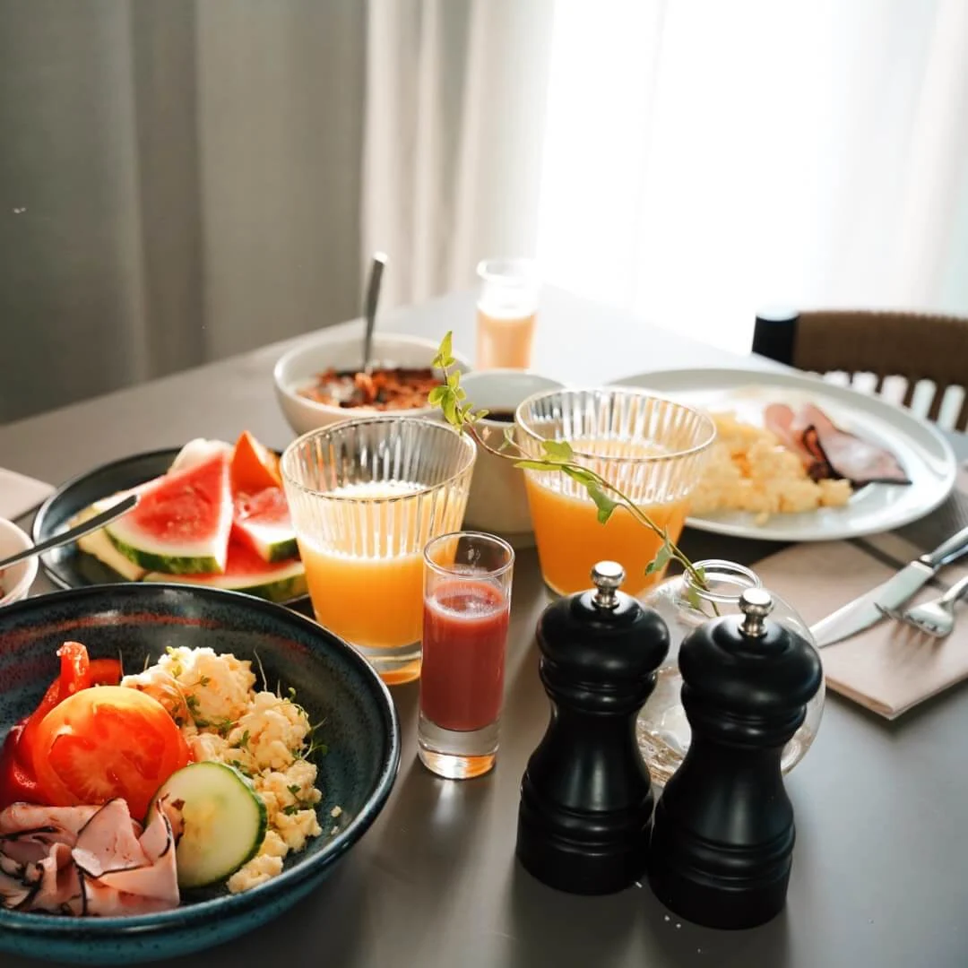 A variety of breakfast foods including bowls of cereal, sliced meats, vegetables, fruit, and drinks are set on a dining table with utensils and napkins on the side.