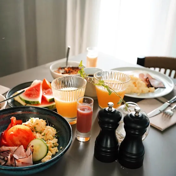 Et variert frokostbord med skåler med frokostblanding, skiver av kjøtt, grønnsaker, frukt og drikker på et spisebord med bestikk og servietter ved siden av.