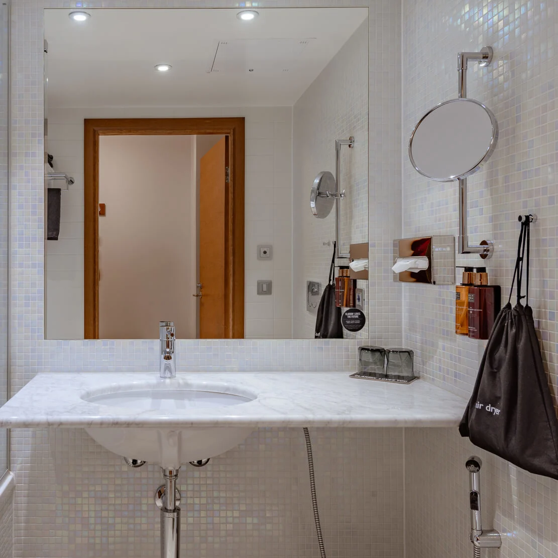 Et moderne badeværelse med en hvid vask under et stort spejl, der reflekterer en dør, omgivet af beige fliser, en bruser og forskellige toiletartikler inklusive en sort pose mærket "air dryer."