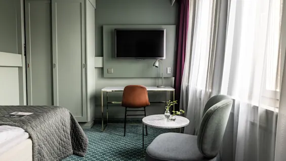 Ett mysigt hotellrum i dämpade gröna toner med en prydligt bäddad säng, skrivbord med lampa och en bekväm stol vid ett runt bord med en vas. Solljus sipprar in genom skira gardiner och skapar en varm atmosfär.