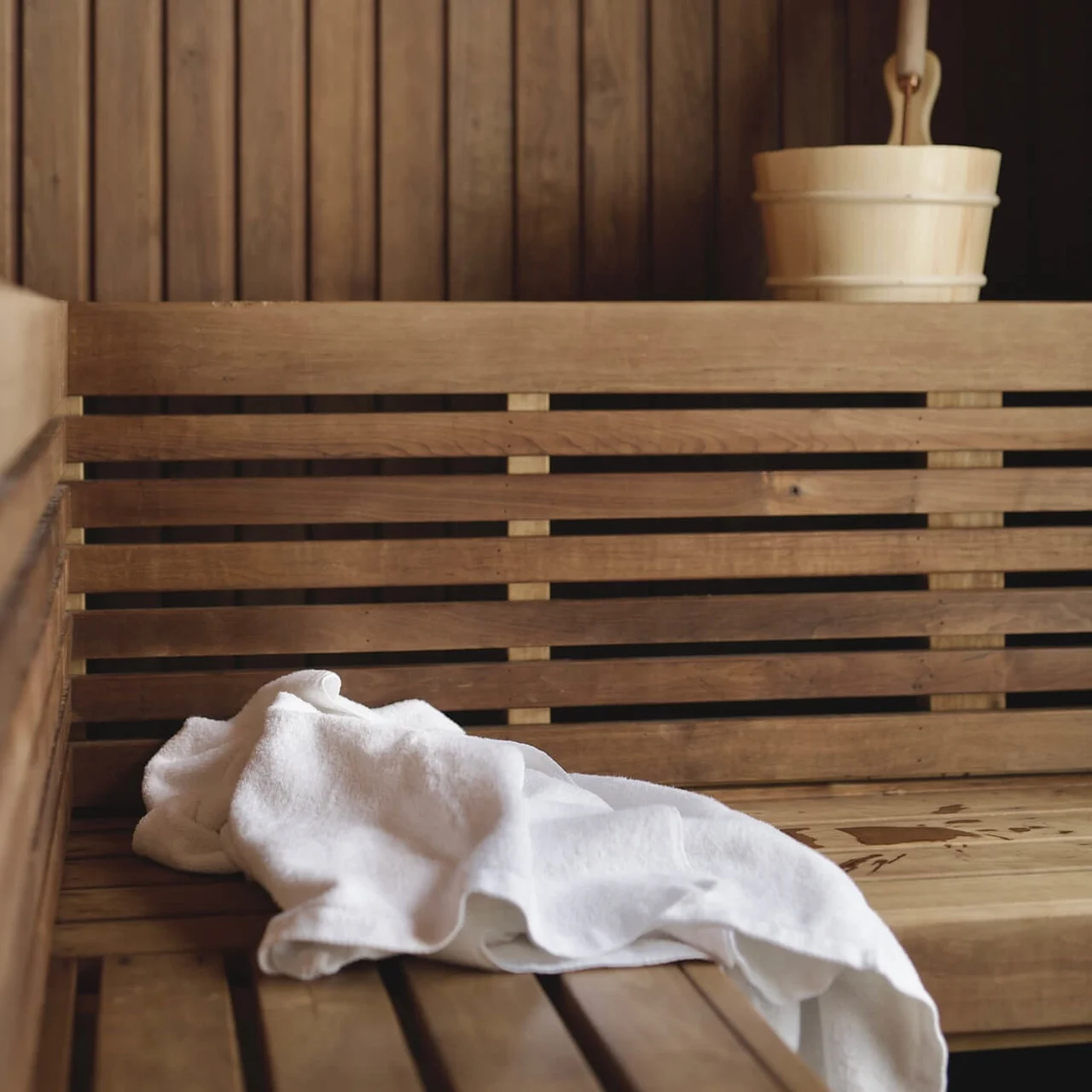 A wooden sauna bench with a white towel, wooden bucket and ladle, symbolizing relaxation and heat.