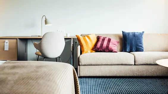 En beige sofa med farverige puder stor i et rum med et mønstret gulv. Ved siden af er et moderne skrivebord med lampe og stol mod en lys væg.