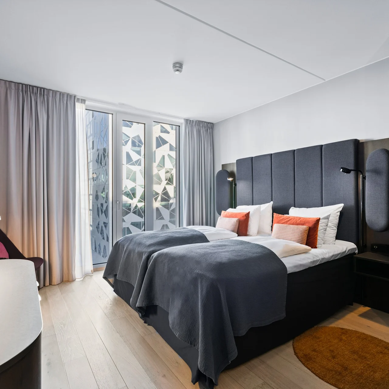 A bed covered with gray blankets and orange pillows is in a modern bedroom. Soft lighting and patterned curtains decorate the room, with furniture including a desk and chair.