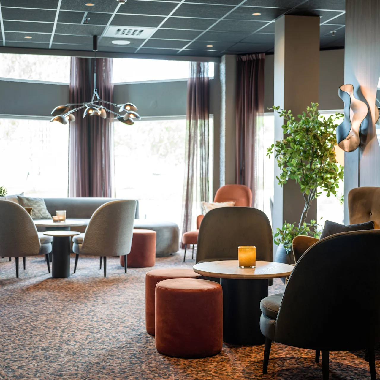Cozy lounge with modern chairs and tables, centered around small candles. Elegant chandelier hangs above, with large windows and a potted plant adding natural light and greenery.