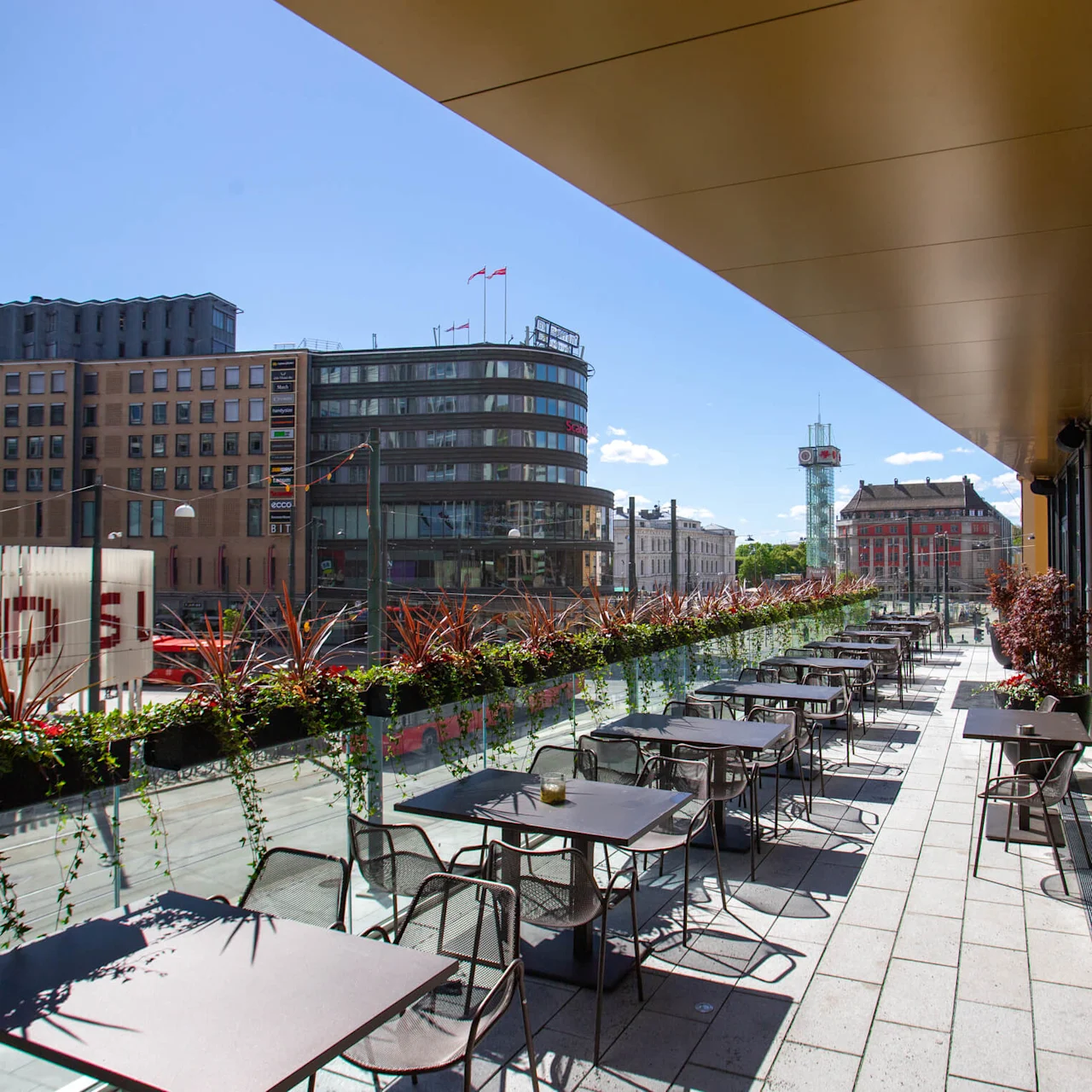 Outdoor restaurant terrace features empty tables and chairs lined with greenery. Positioned above a bustling urban street, surrounded by modern buildings under a clear blue sky.