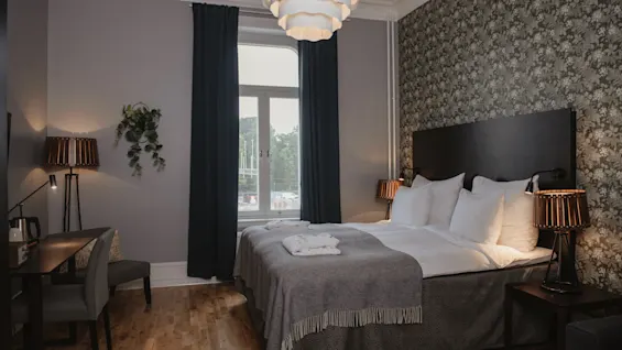 A comfortable hotel room features a neatly made bed with white linens against a patterned wall. A desk and chair sit nearby under soft lighting. A window offers an outside view.