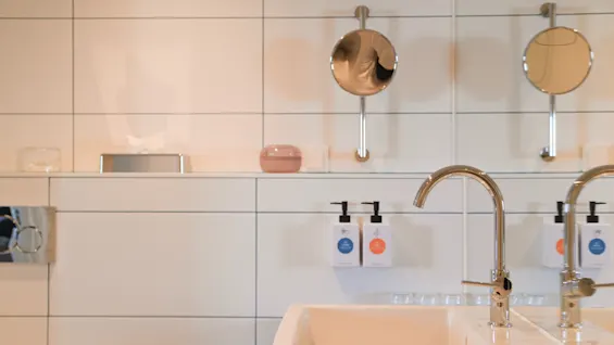 To hvide håndvaske med kromhaner; over dem, dispensere med forskellige farvede etiketter giver sæbe. I baggrunden en hvid flisevæg med to cirkulære spejle og små toiletartikler.