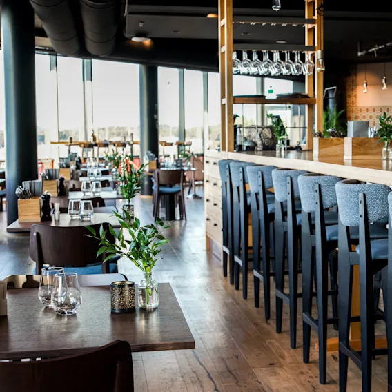 En moderne restaurantinteriør har en lang bar med høye stoler, opplyst av hengelamper. Bord med vaser og glass fyller rommet med tregulv, store vinduer slipper inn naturlig lys.