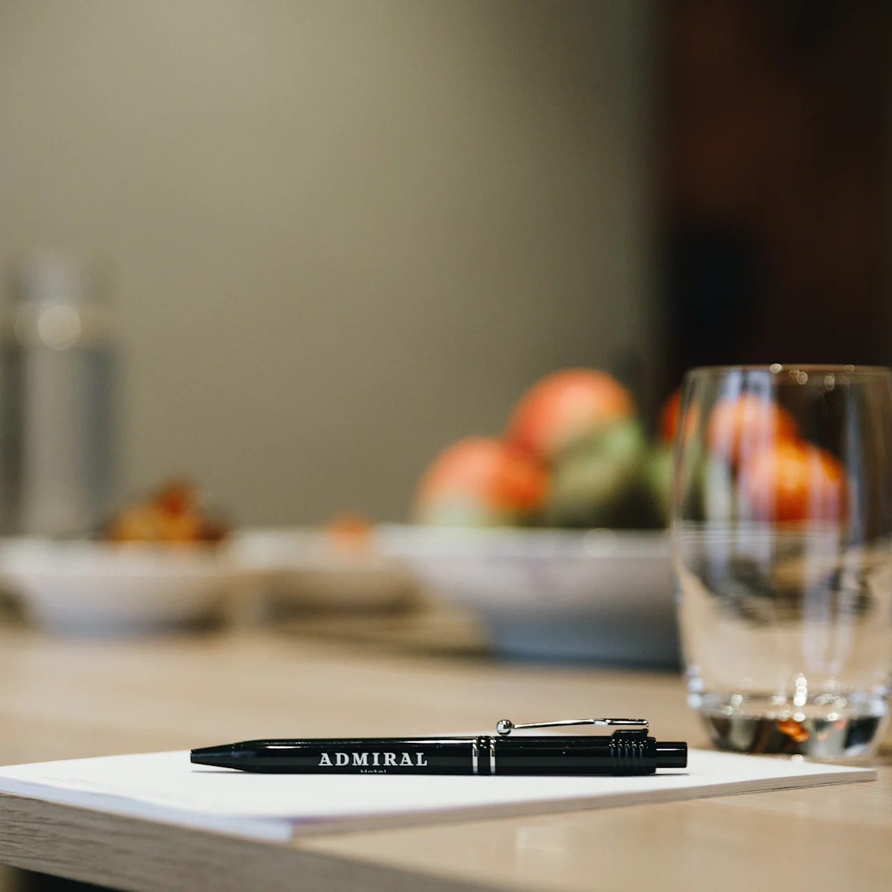 Pen labeled "ADMIRAL" resting on a closed notebook; next to a clear glass on a wooden table in a room with blurred bowls of fruit in the background.