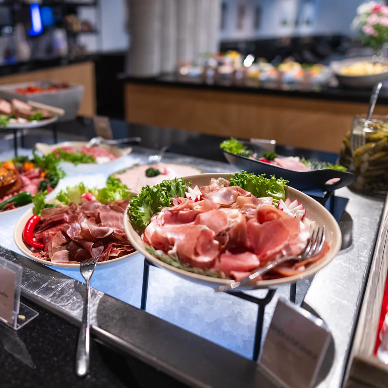 Tallerkener fyldt med forskellige skiver af kød, pyntet med grønt, fremvises på en buffet med serveringsbestik. Nærliggende skilte siger "FORELØB," "SYRNET OST," og "MORGON BUFFÉ."