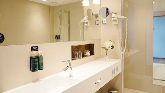 A modern bathroom with a sleek countertop holding a faucet, toiletries, and a flower vase beside a large mirror. Wall-mounted bottles labeled "hand & body wash." Overhead lights illuminate the space.