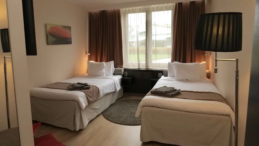 Två sängar är prydligt arrangerade under ett stort fönster med dragna bruna gardiner, i ett mysigt hotellrum med trägolv och en väggmonterad målning.
